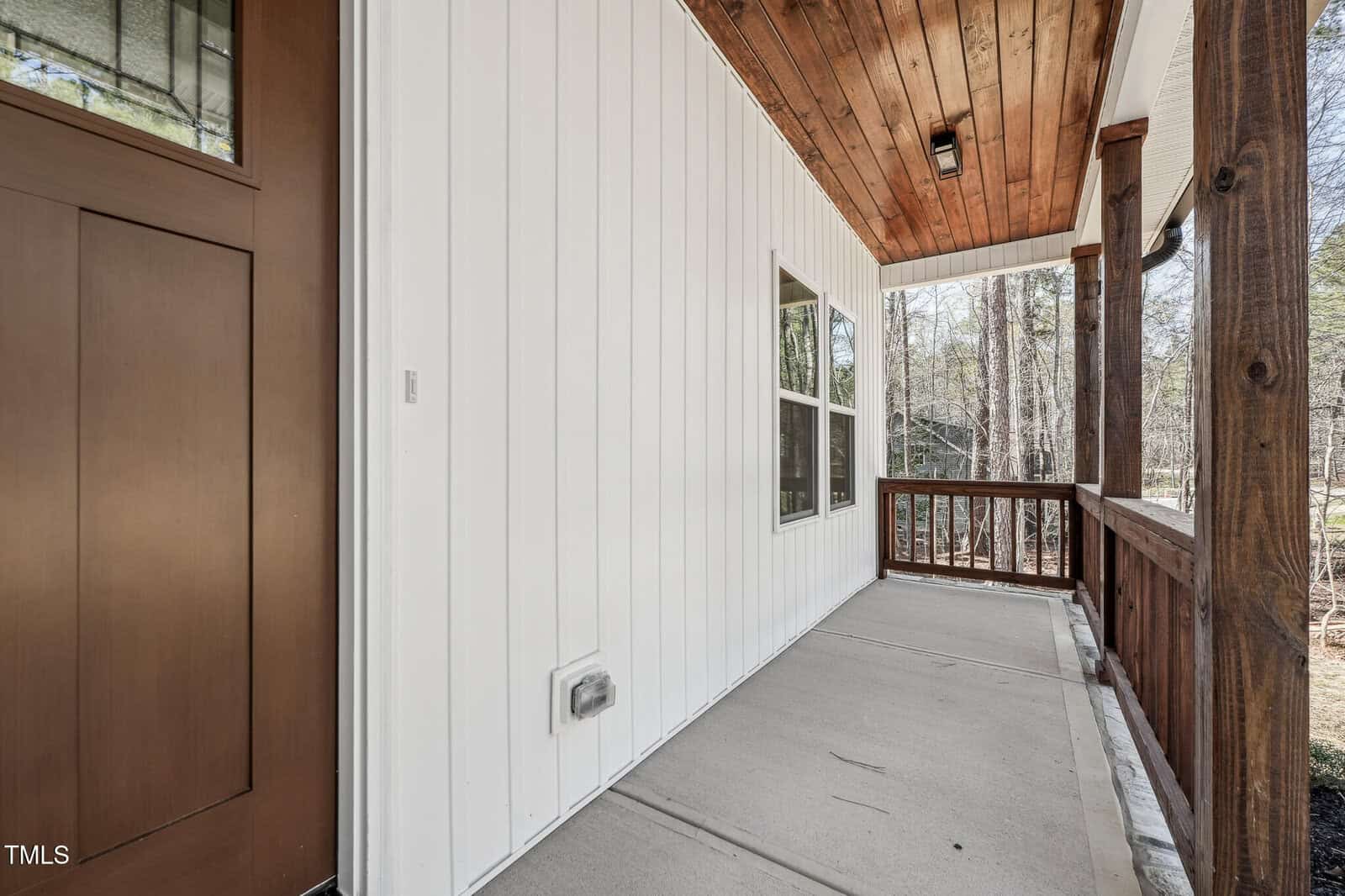 A covered porch with a wood ceiling and railing, white vertical siding, a brown door on the left, and a window facing the porch. The floor is concrete, and trees are visible in the background.