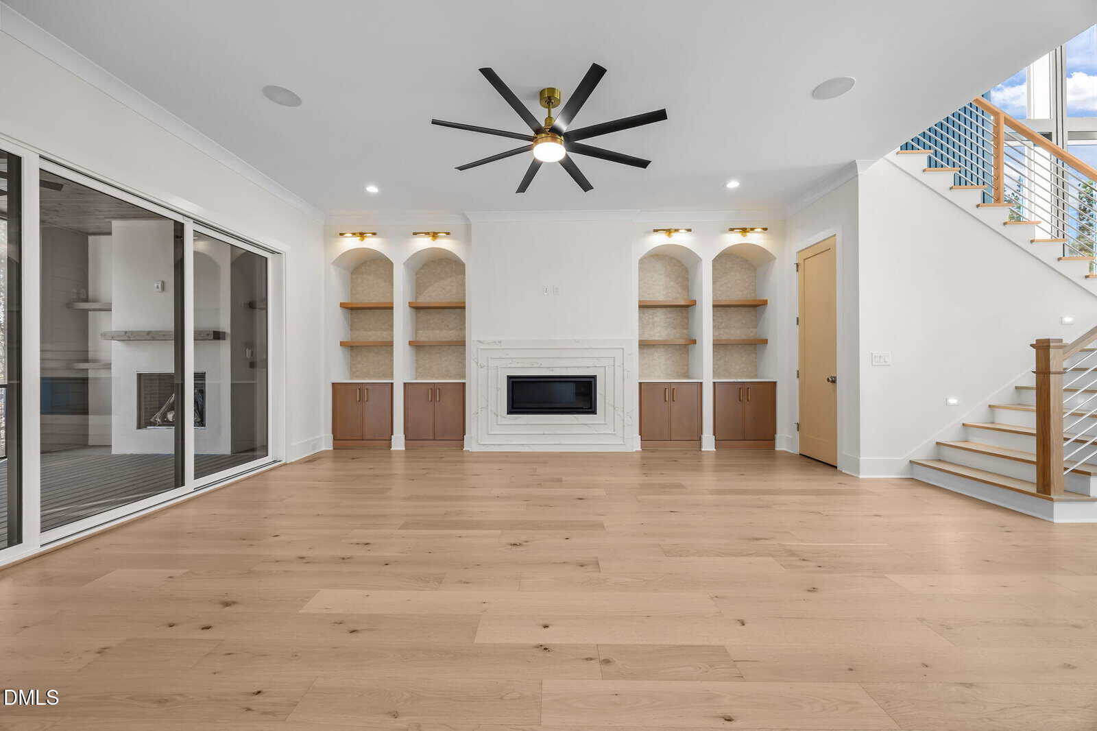 Modern living room with light wood floors, built-in shelves and cabinets, a central fireplace, a black ceiling fan, and glass doors leading to another room. Stairs with a blue railing are visible on the right.