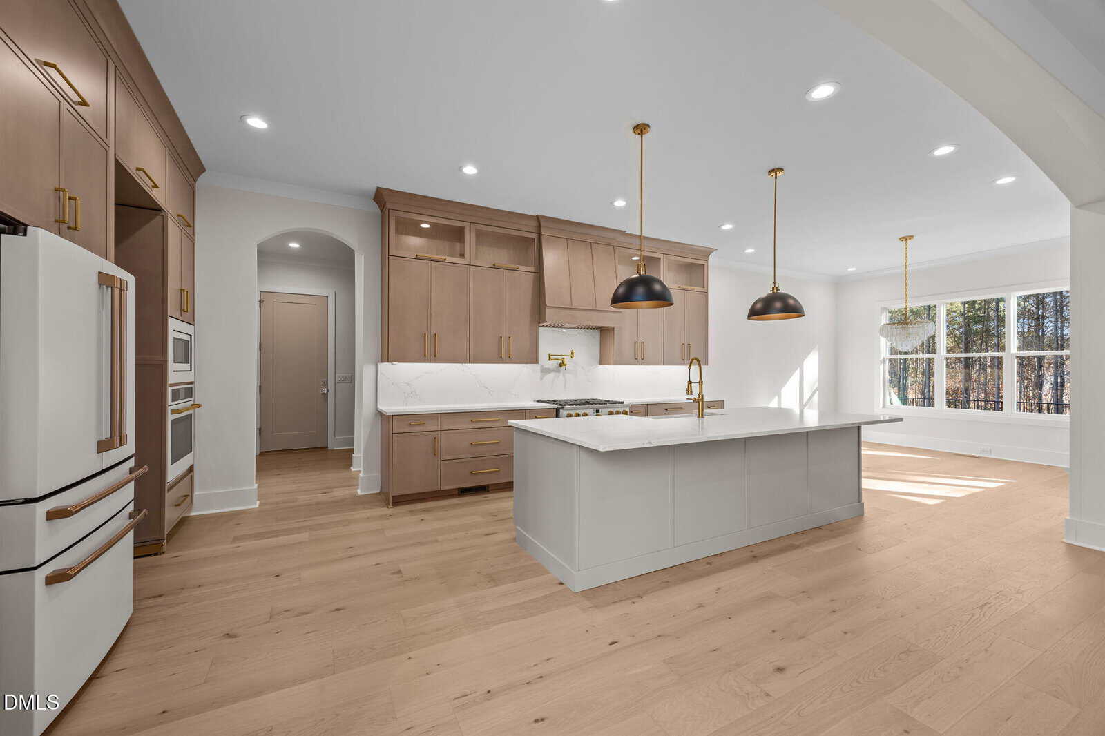 Modern kitchen with light wood floors, tan cabinets, a white island with gold accents, white appliances, two black pendant lights above the island, and large windows providing natural light in the dining area.