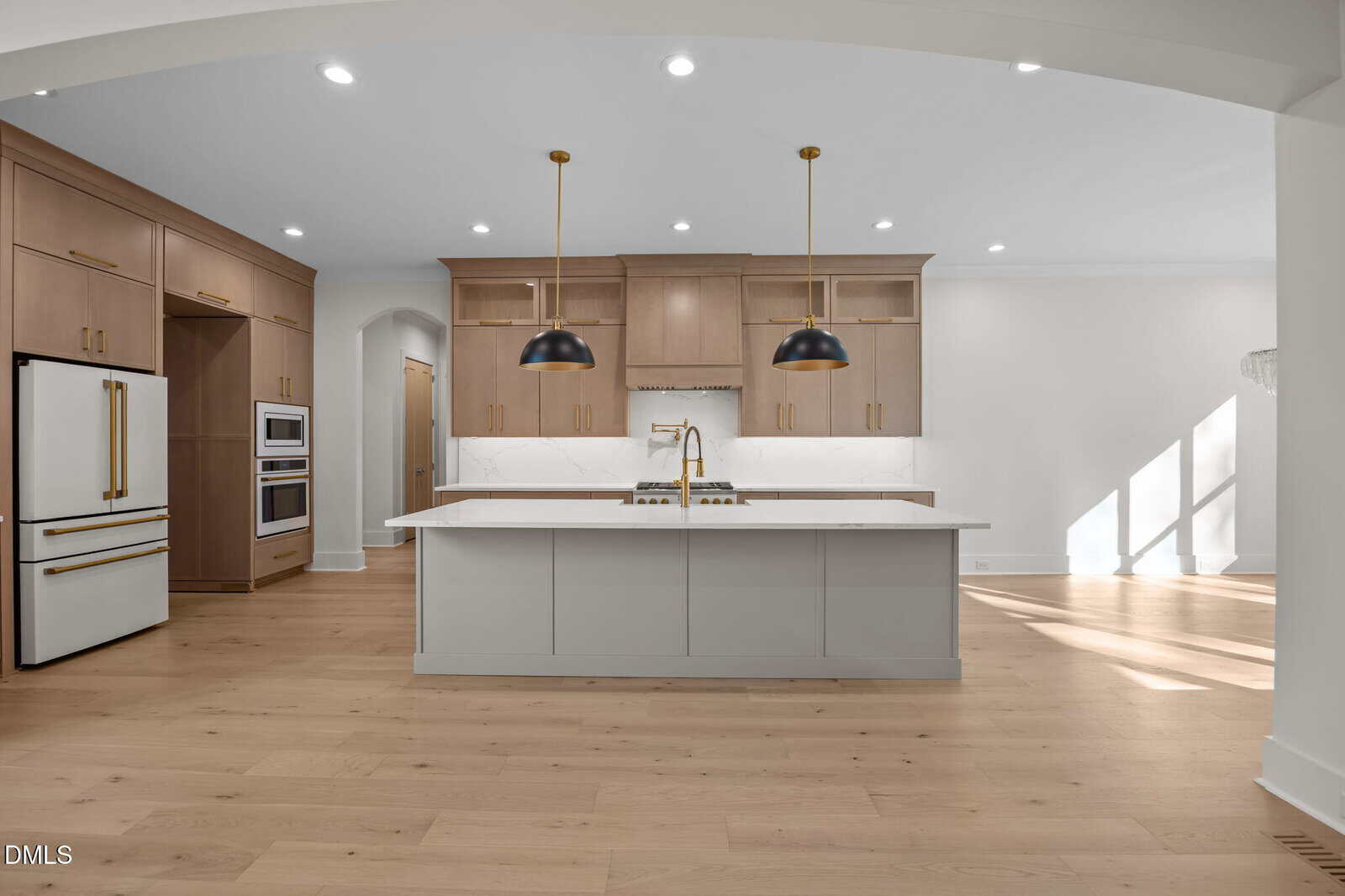 Modern kitchen with light wood floors, beige cabinets, a white island with gold hardware, pendant lights above the island, built-in oven and refrigerator, and white marble backsplash and countertops.