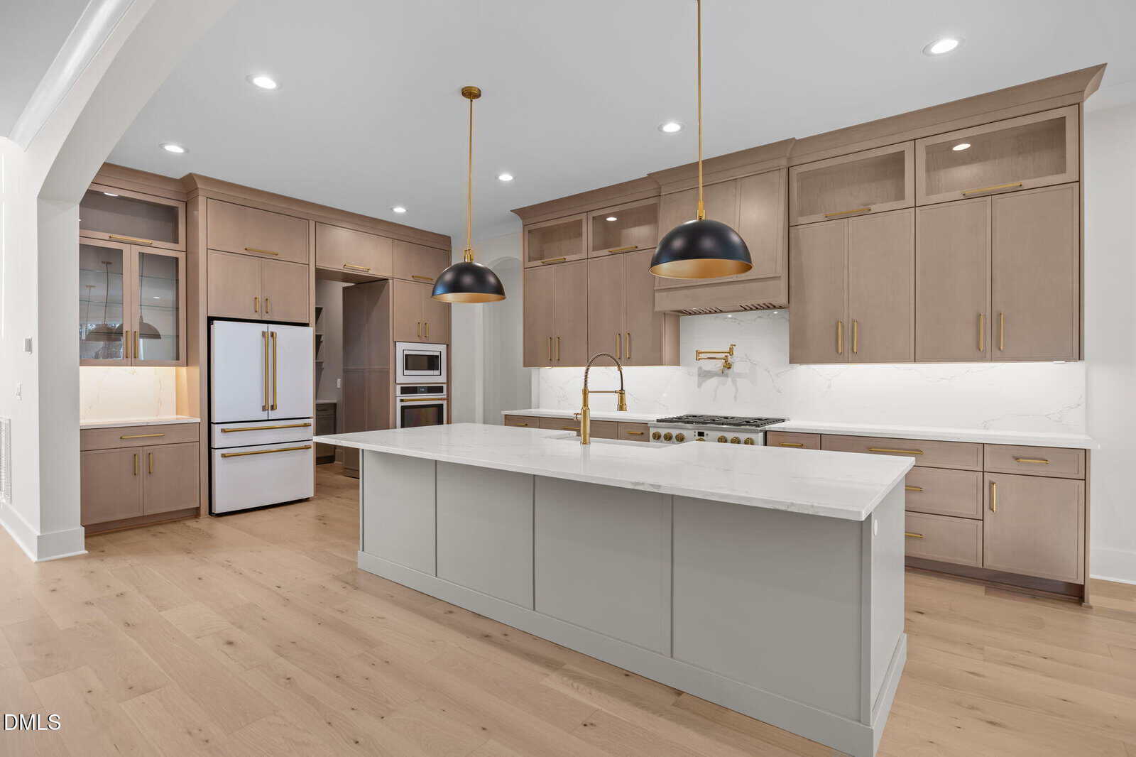 Modern kitchen with light wood floors, taupe cabinets, a large center island with a white countertop, gold hardware, pendant lights, built-in appliances, and a white refrigerator.