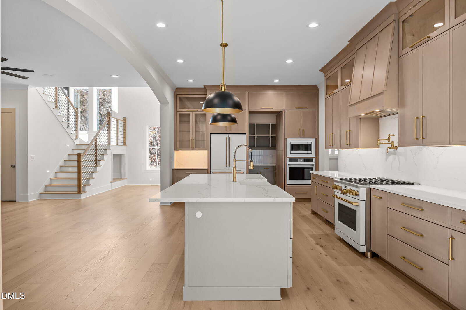 Modern kitchen with light wood floors, beige cabinets, white marble countertops, gold fixtures, stainless steel appliances, large island with sink, and pendant lighting. Open layout with a staircase visible in the background.