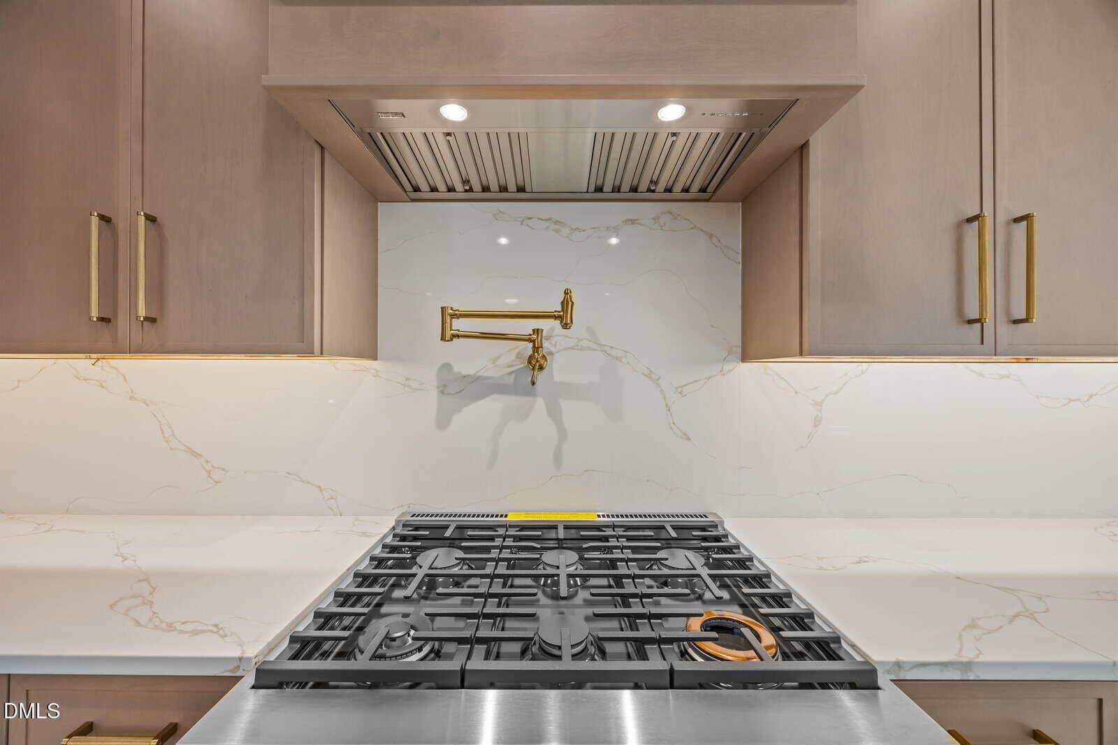 A modern kitchen with a stainless steel gas stove, marble backsplash, gold pot filler faucet, and light wood cabinets with gold handles.