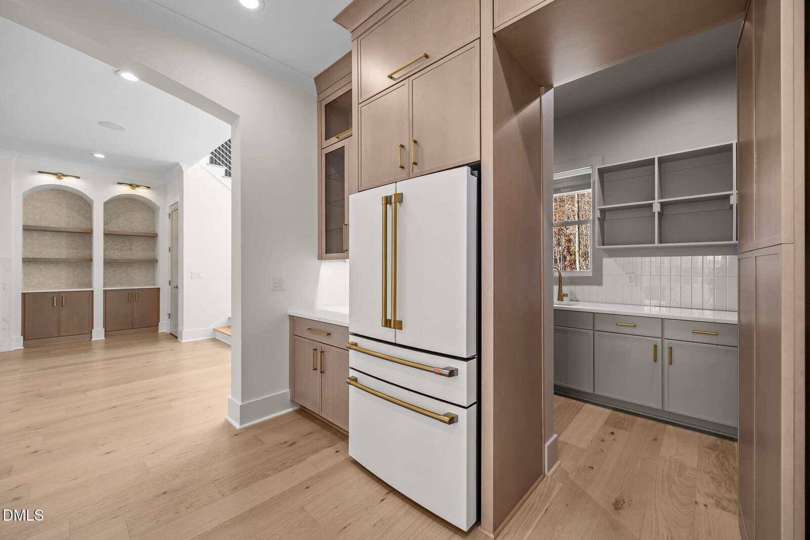 Modern kitchen with light wood floors, white and beige cabinets, a white refrigerator with gold handles, and a view into a pantry area with gray cabinets, a window, and white tile backsplash. Built-in shelves visible in hallway.