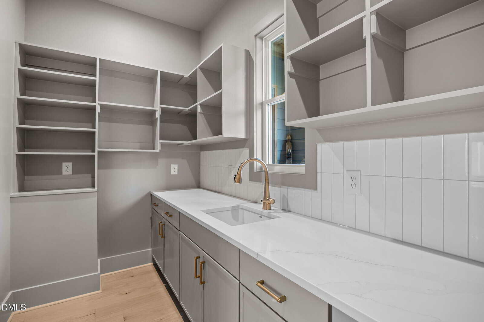 Modern laundry room with light gray cabinets and open shelves, a long white countertop with a built-in sink, gold hardware, white tile backsplash, and a window above the sink. The floor is light wood.