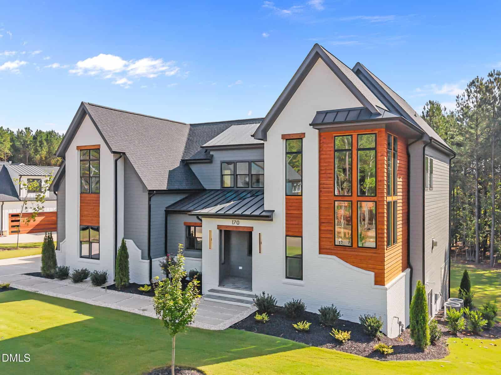 A modern two-story house with large windows, white and gray exterior walls, and wooden accents. The home is surrounded by a manicured lawn, small shrubs, and a backdrop of tall trees under a blue sky.