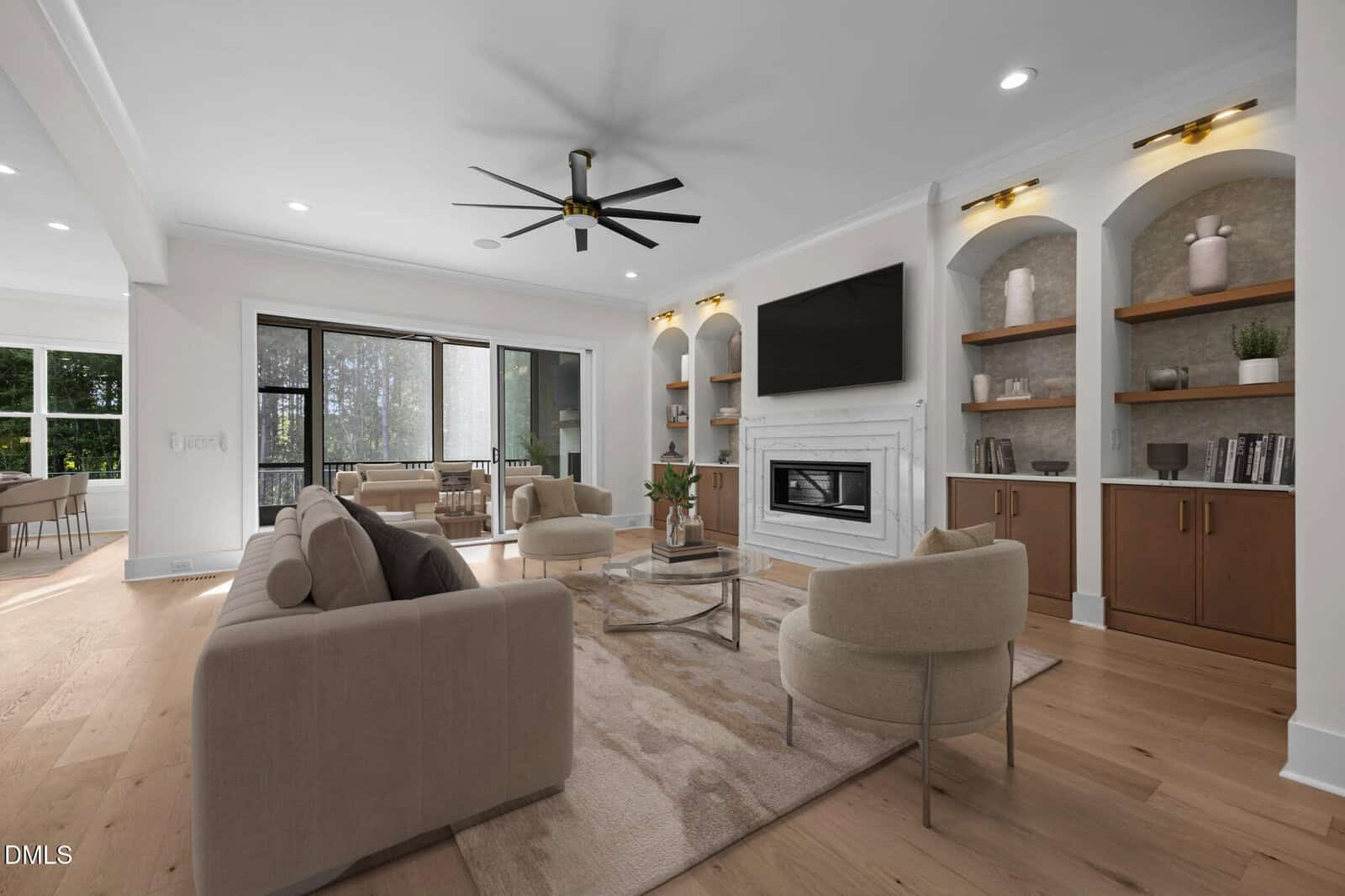 Modern living room with beige sofas, two round chairs, a glass coffee table, fireplace, wall-mounted TV, built-in shelves with decor, light wood flooring, large windows, and ceiling fan. Adjoining sunroom visible in the background.