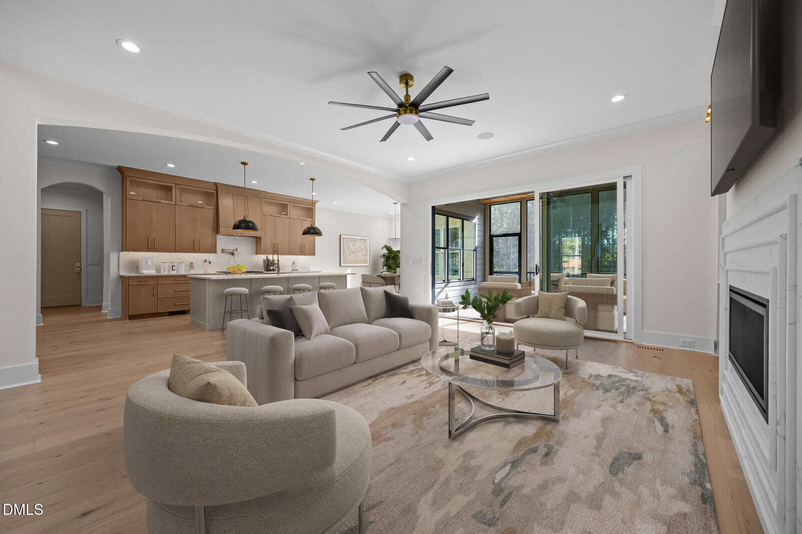 A modern open-concept living room and kitchen with neutral-toned furniture, wood floors, a round glass coffee table, and large windows leading to a sunroom with outdoor views. A ceiling fan hangs above the seating area.