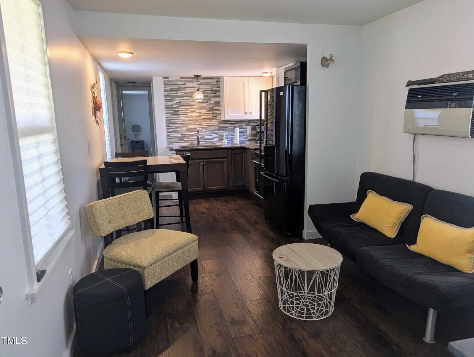 A small living space with a dark floor features a couch with black cushions, a yellow patterned chair, a round white wireframe coffee table, and a dining area with high chairs. The kitchen has a tile backsplash, dark cabinets, and a black refrigerator.