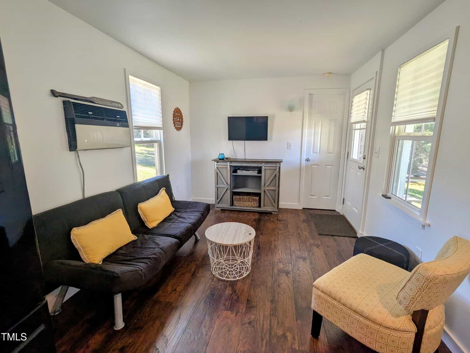 A small living room with a black futon adorned with yellow cushions, an armchair, and a white round table. A flat-screen TV is mounted on the wall above a console. Two windows and a window-mounted air conditioner are visible. Wood flooring throughout.