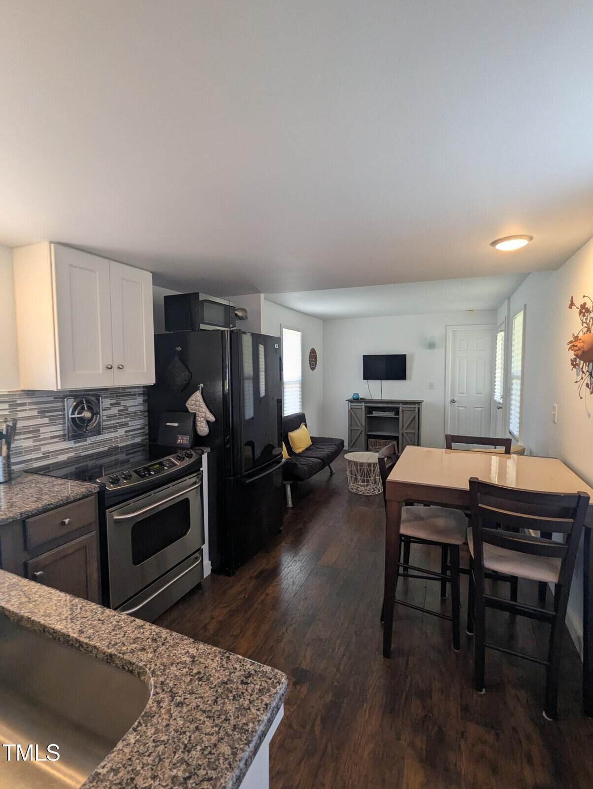 Modern kitchen and living area with granite countertops, stainless steel stove, black refrigerator, and white cabinets. There's a dark wood dining table with chairs, a small couch, and a wall-mounted TV. The room has wooden flooring and a baseboard heater.