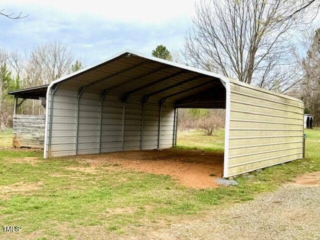 A gray metal carport with an open front stands on a patch of bare earth and grass. Nearby, there is a smaller wooden structure. The background features leafless trees and a cloudy sky.