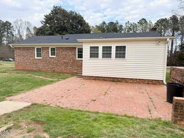 A single-story house with a brick exterior and a white siding extension. It has a large, paved patio area and is surrounded by grass. Trees and a cloudy sky are in the background.
