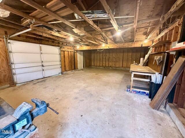 A spacious, empty workshop with a wooden workbench on the right, a metal vice on a table in the foreground, and a partially open garage door on the left. Exposed wooden beams are visible on the ceiling.