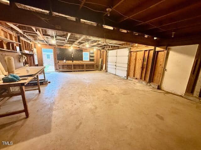 A spacious garage interior with wooden beams and a concrete floor. There is a workbench on the left, a closed white garage door on the right, and a door leading outside in the background. The walls are partially unfinished with exposed wood.