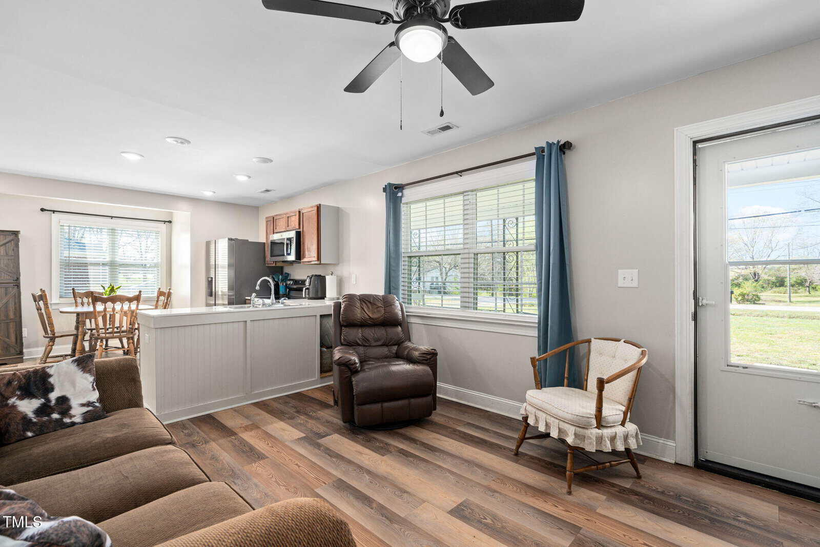 A living room with wood flooring, brown sofa, recliner, white chair, and a ceiling fan. The kitchen with stainless steel appliances and a dining area are visible in the background. Sunlight comes through large windows.