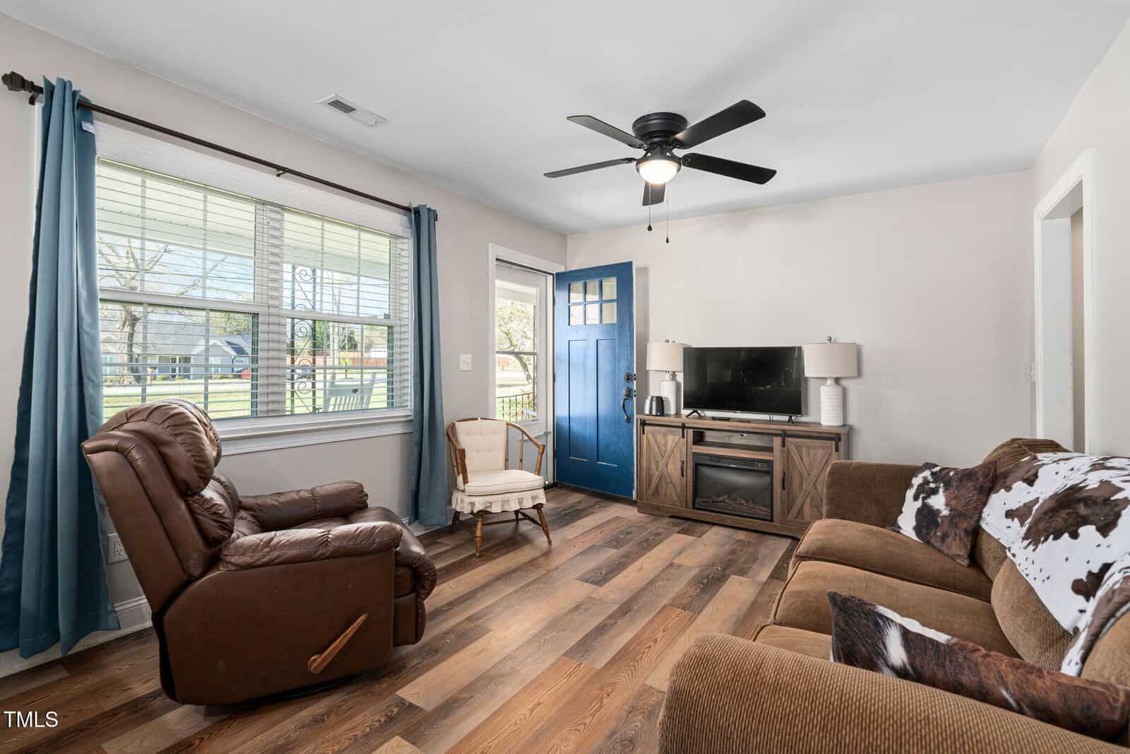 Living room with wood flooring, brown sofa, leather recliner, accent chair, TV on a stand, ceiling fan, blue front door, and two windows with blue curtains. Walls are light-colored and natural light enters from the windows.