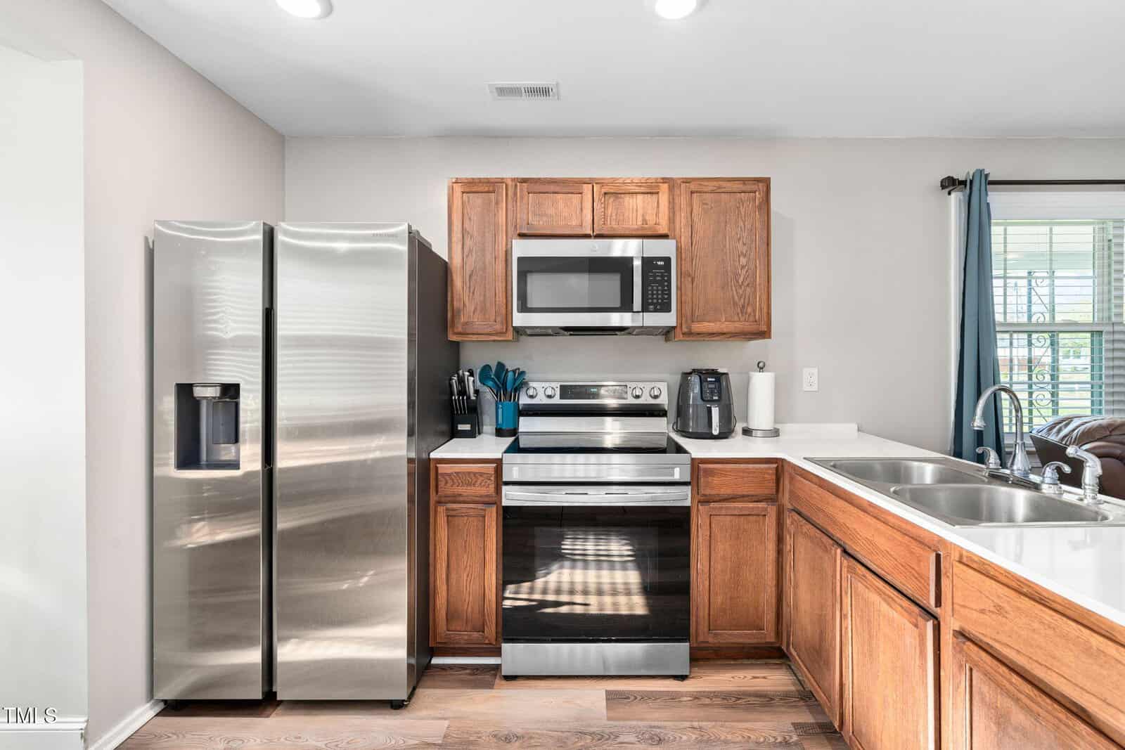 A kitchen with stainless steel refrigerator, electric stove, and microwave, brown wooden cabinets, white countertops, a sink, and a window with blue curtains. Utensils are on the counter next to the stove.