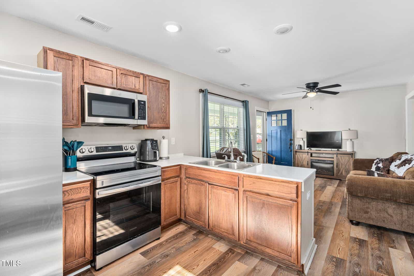A kitchen with wooden cabinets, stainless steel appliances, and white countertops opens into a living room with a brown sofa, television, and a blue front door. Light wood flooring runs throughout the space.
