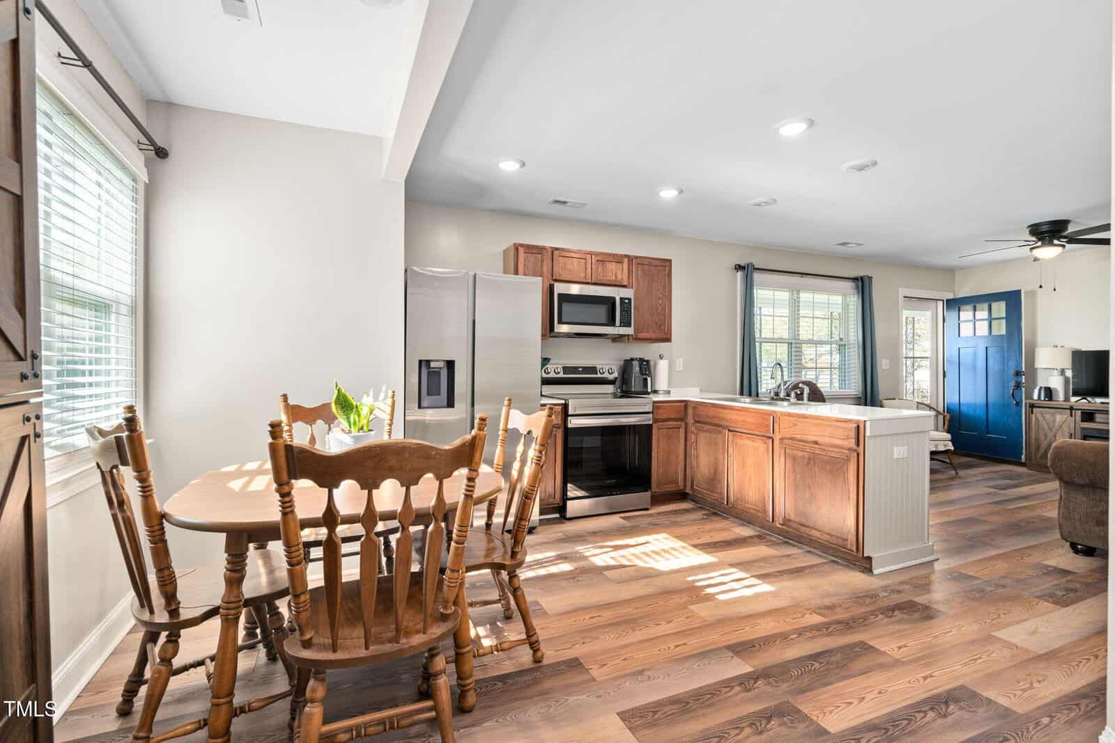 A kitchen and dining area with wooden floors, a wooden dining table and chairs, stainless steel appliances, wooden cabinets, a kitchen island, and a blue front door visible in the background.