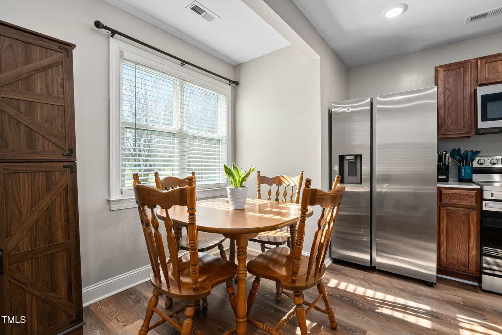A small wooden dining table with four matching chairs sits by a window with blinds. A potted plant is centered on the table. Stainless steel fridge, stove, and cabinets are visible in the adjacent kitchen.