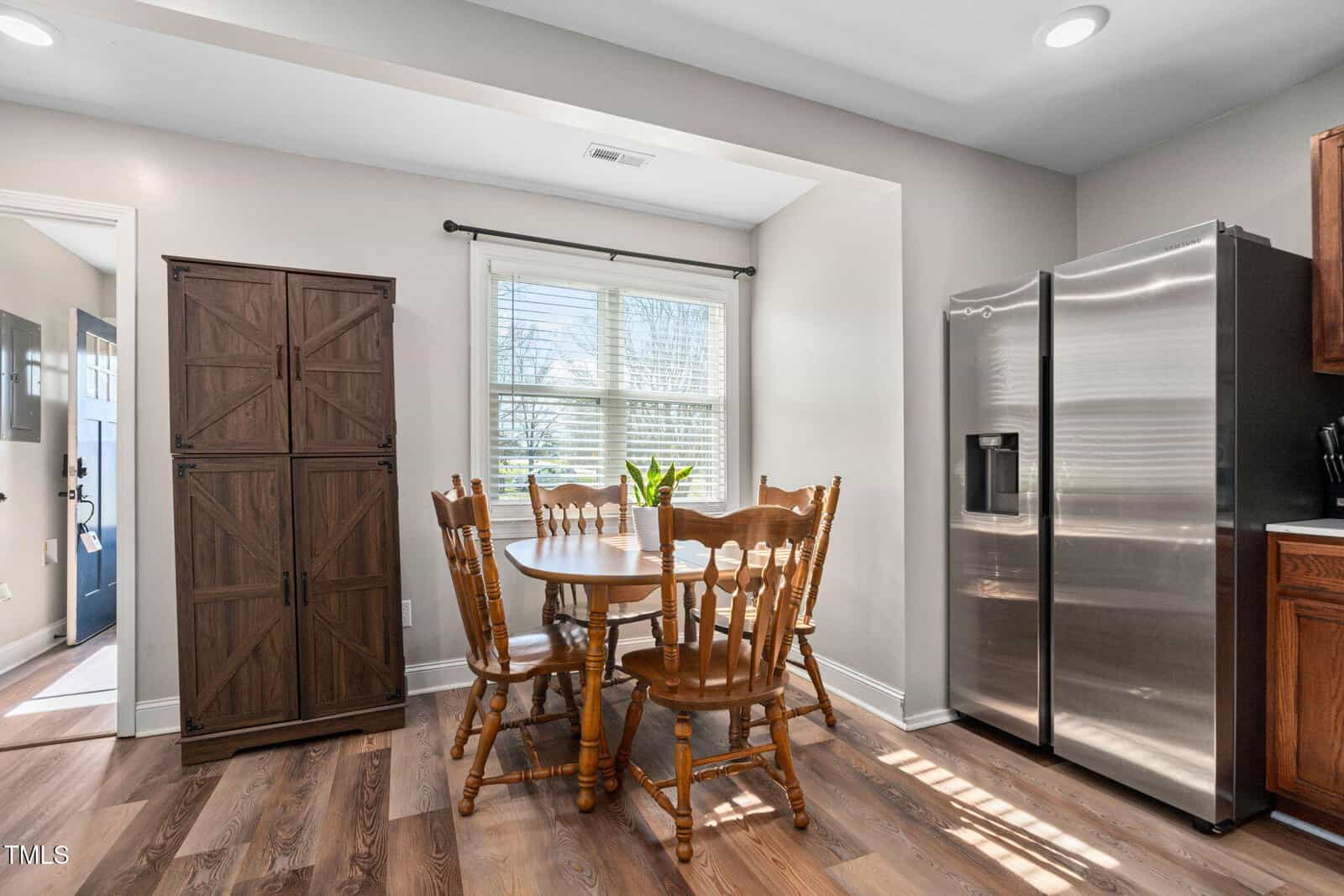 A small dining area with a round wooden table and five matching chairs is next to a window. A tall cabinet stands to the left, and a stainless steel refrigerator is on the right. Sunlight enters through the window.