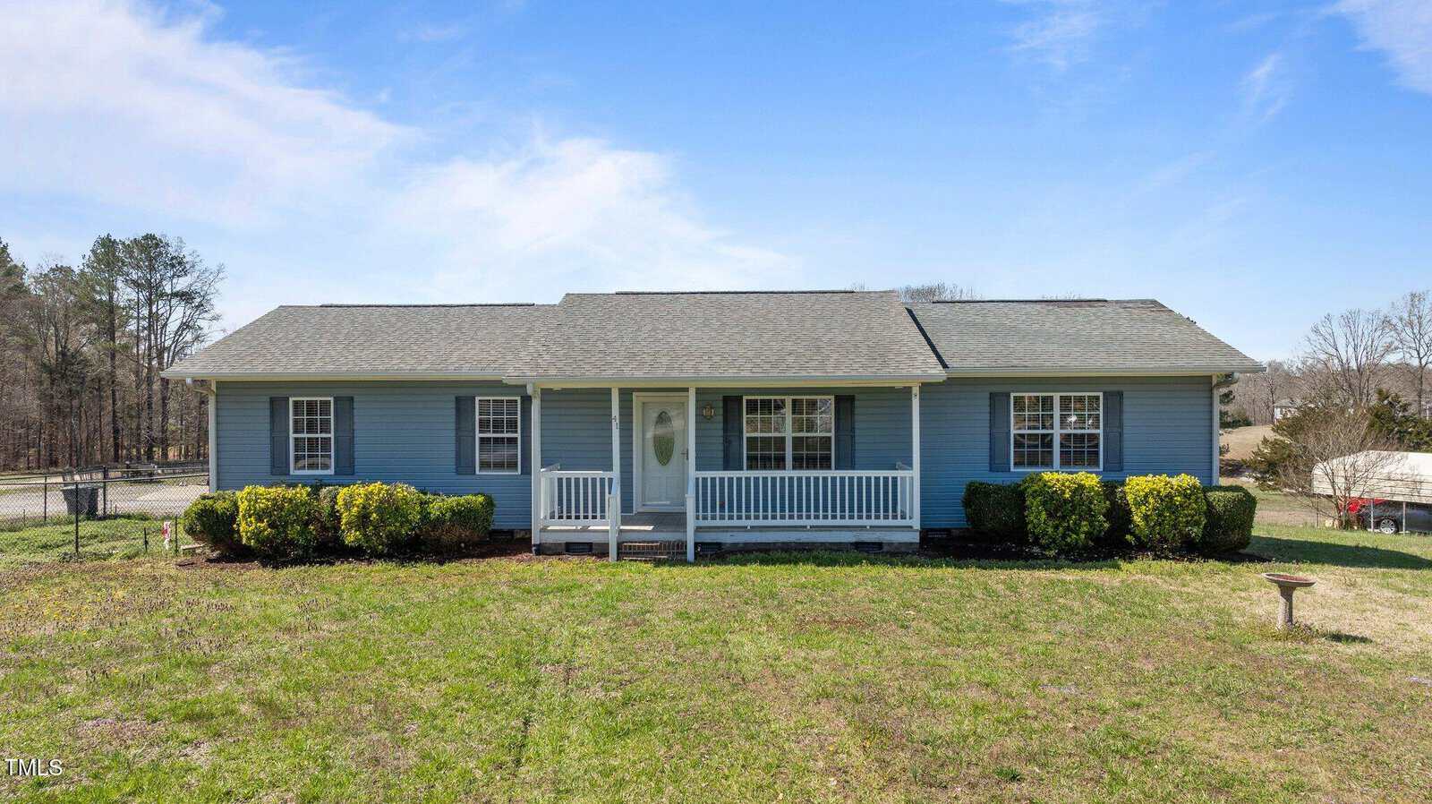 Nestled at 41 Rock Bluff Lane in Kittrell, this charming single-story house boasts blue siding and a gray roof. A cozy front porch with white railings invites you in, overlooking a grassy yard dotted with bushes. The scene is complete with a clear, sunny sky as its backdrop.