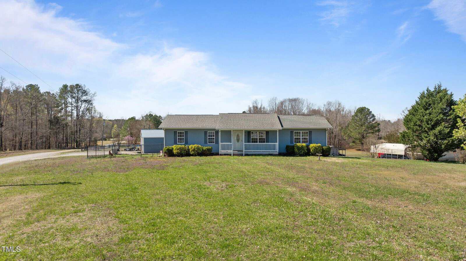 A small, single-story blue house with a covered front porch and a neatly trimmed lawn. The house is surrounded by sparse trees, and a road runs nearby under a clear blue sky.