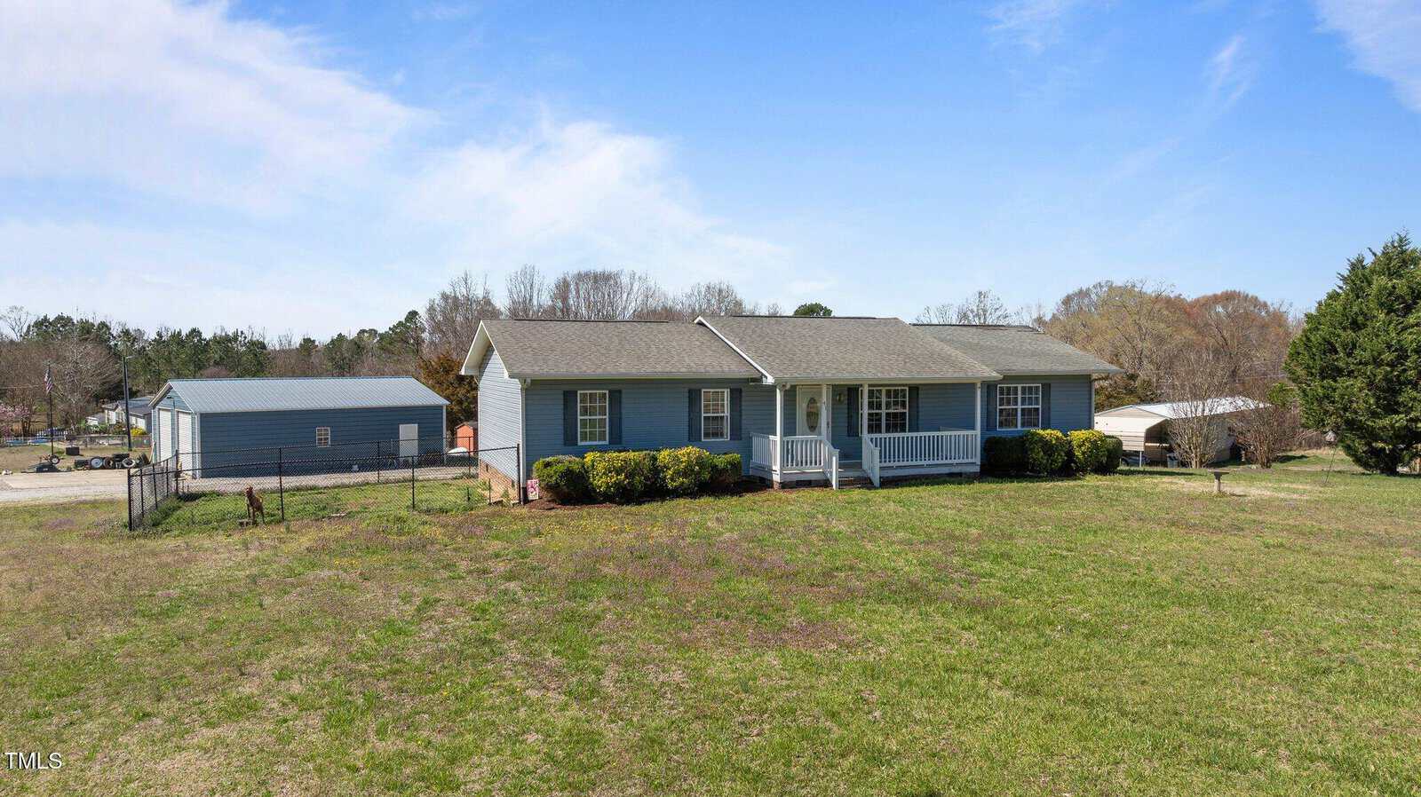 A single-story house with a light gray exterior and a covered front porch is situated on a large grassy lawn. A fence and a detached garage or shed are visible in the background. The sky is blue with scattered clouds.
