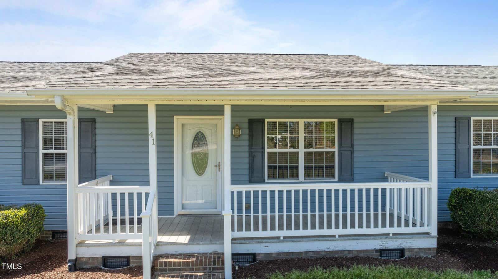 A single-story house with light blue siding and a gray shingled roof. It features a white front door with glass, flanked by two windows with shutters. A white railing frames the small front porch. Trimmed bushes are visible below the porch.