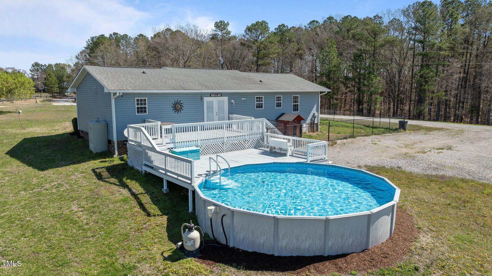 A gray single-story house with a light green roof is shown. In front, a large above-ground pool is surrounded by a white wooden deck. Grass and trees are visible in the background under a clear blue sky.