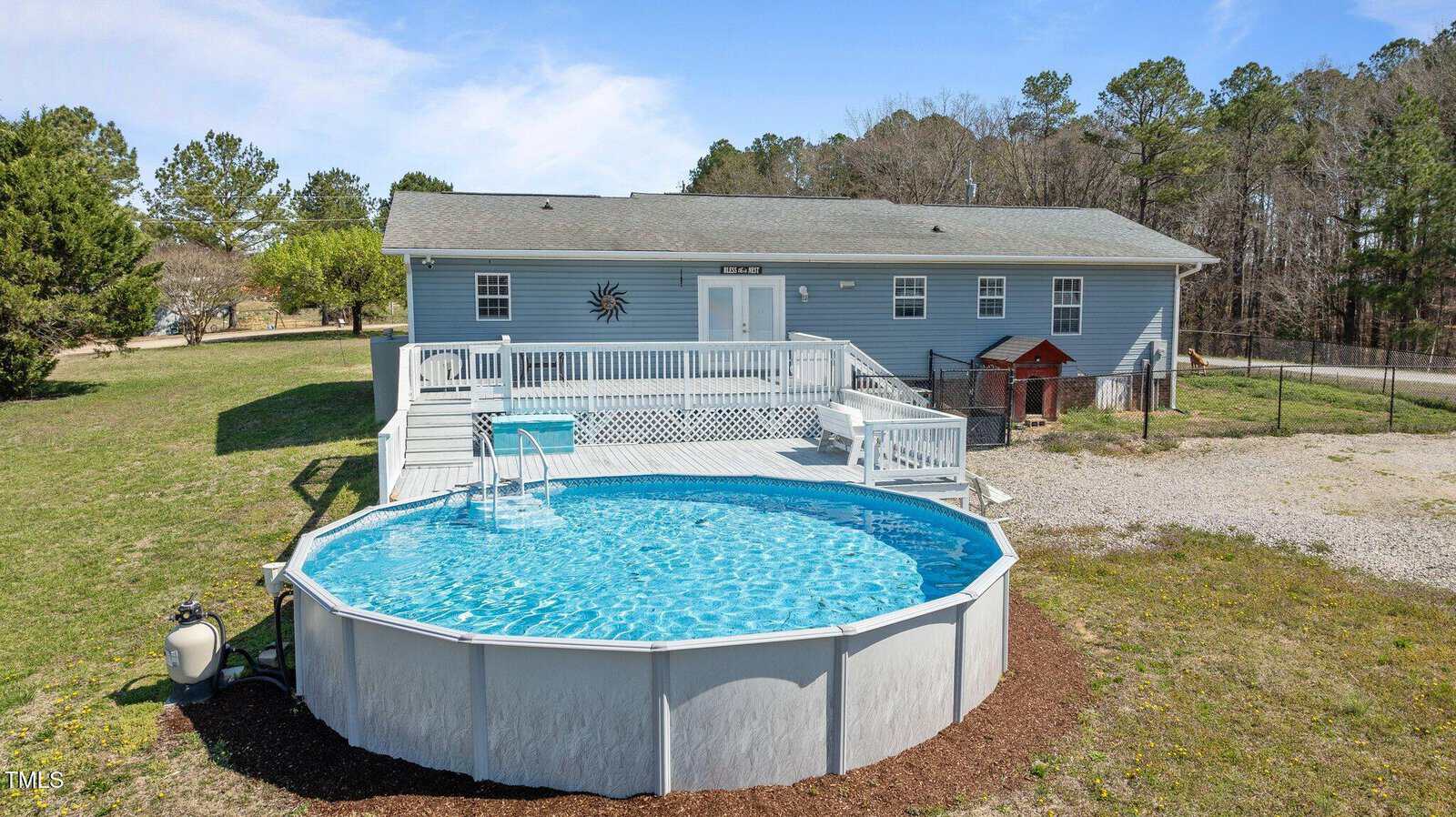 An above-ground round pool filled with blue water is in the foreground, surrounded by a white deck with a few steps. Behind the pool is a single-story light blue house with a gable roof, located in a grassy and wooded area.