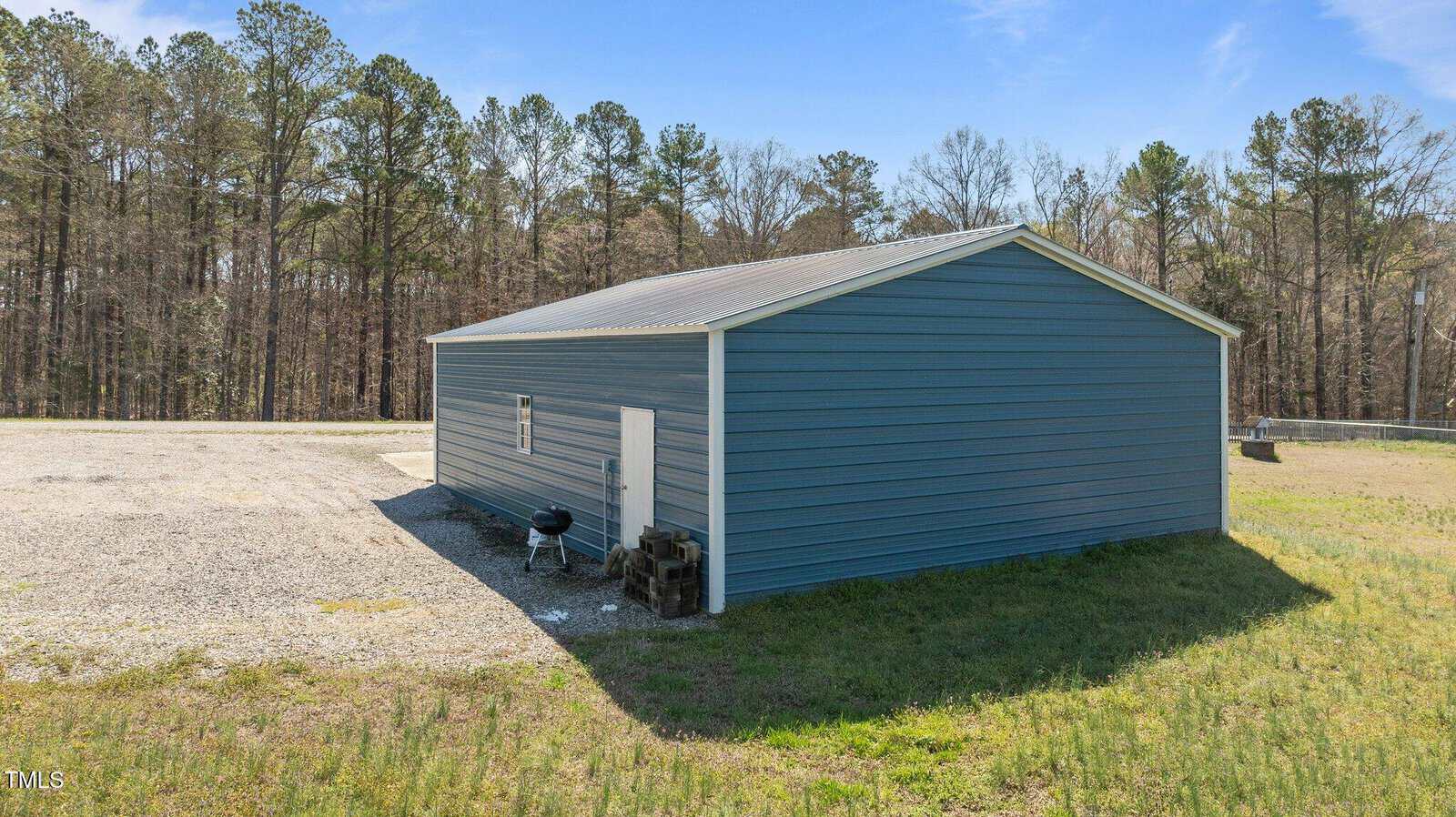 A blue metal building with a pitched roof stands on a gravel surface, surrounded by grass. A small grill and stacked firewood are visible at the side. The background features a wooded area under a clear blue sky.