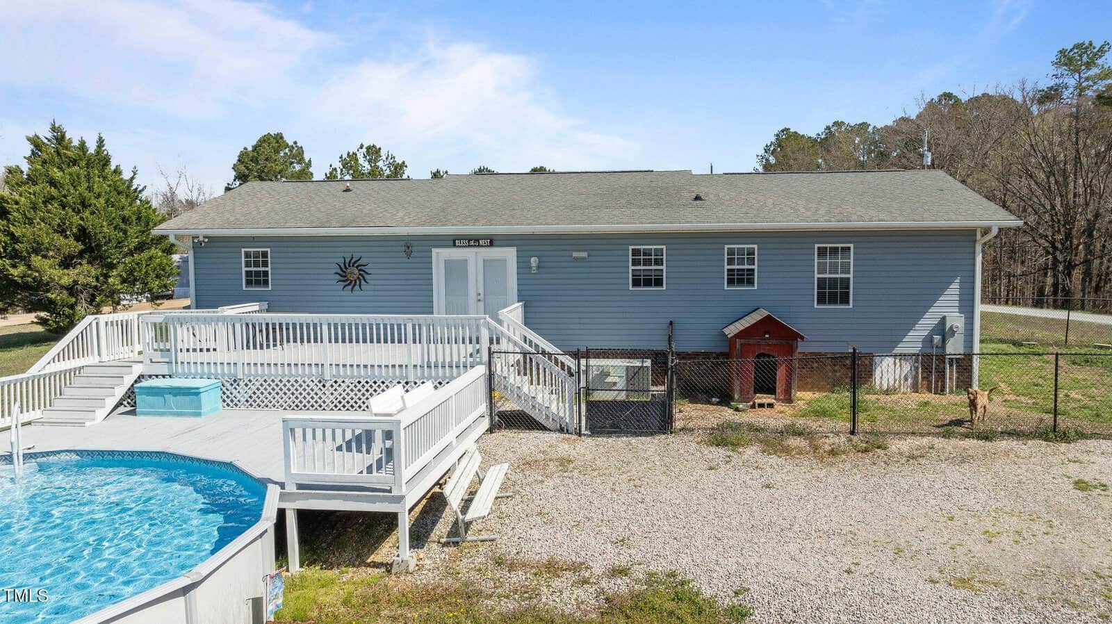 A blue, single-story house with multiple windows and a gray roof sits behind a fenced yard. A round, above-ground pool with a white deck is in the foreground. There's a small red structure and a gravel area next to the fence. Trees are in the background.