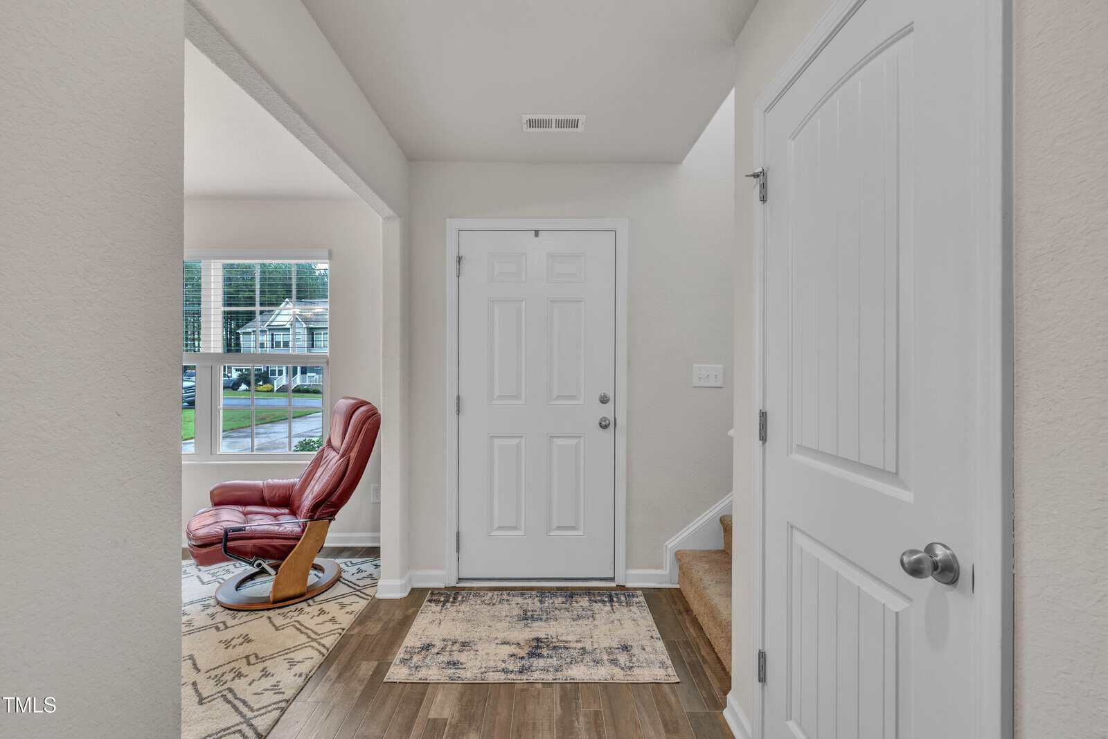 A white front door entryway with a rug, wood flooring, a closed closet door on the right, stairs leading up, and a red recliner chair by a window in an adjacent room.