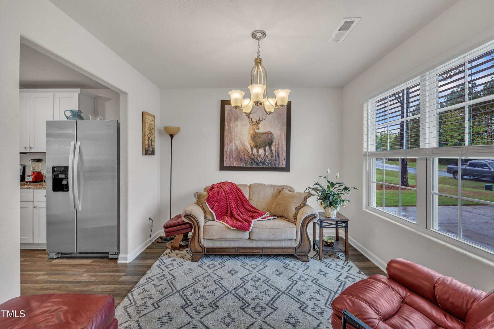 A small living room with a beige loveseat, red blanket, floor lamp, deer painting, and a side table with a plant. The room has wood flooring, a patterned rug, and a window showing a car and grass outside.