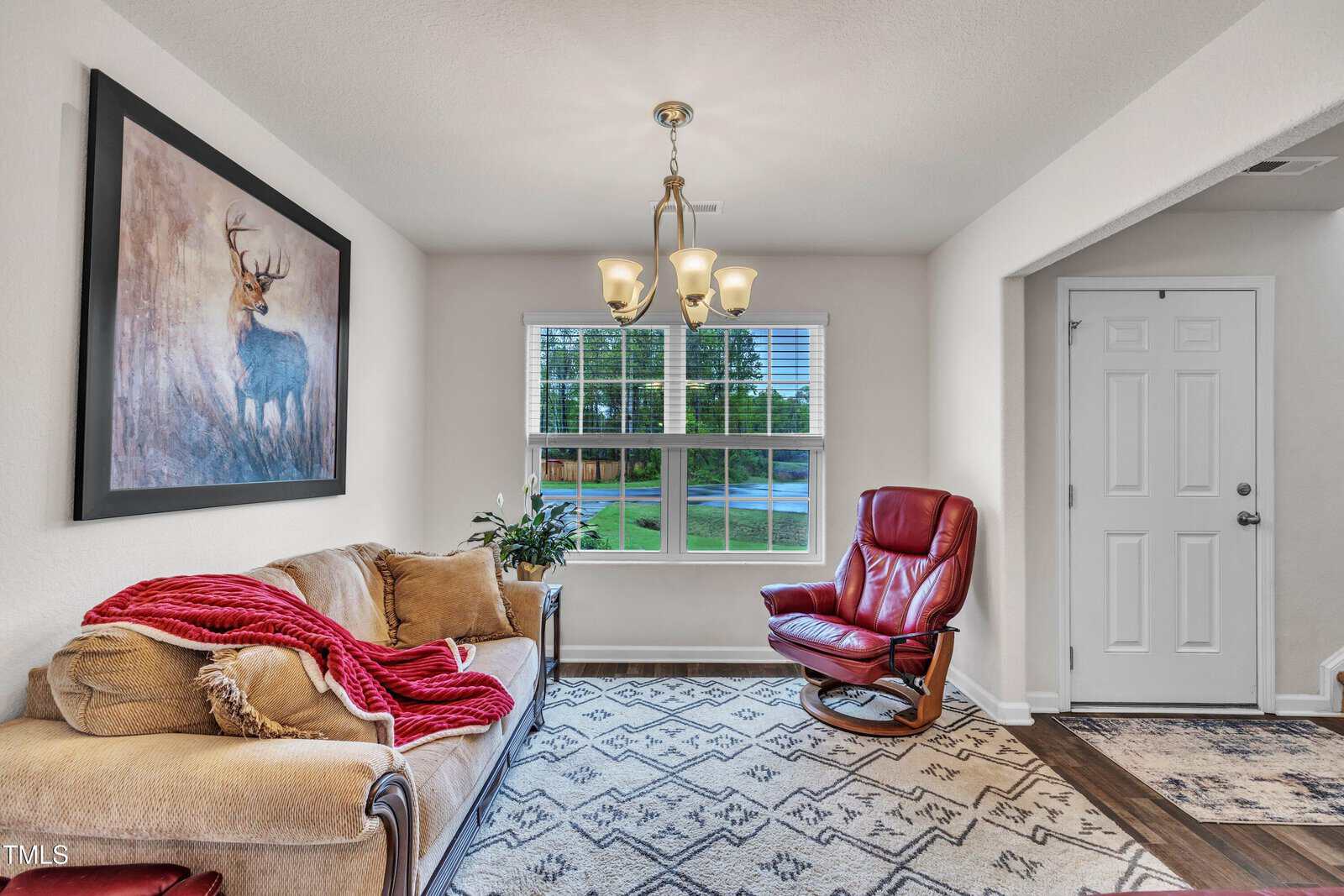 A small living room with a beige couch, red throw blanket, red armchair, patterned rug, large window, chandelier, and deer painting on the wall. The entrance door is visible to the right.