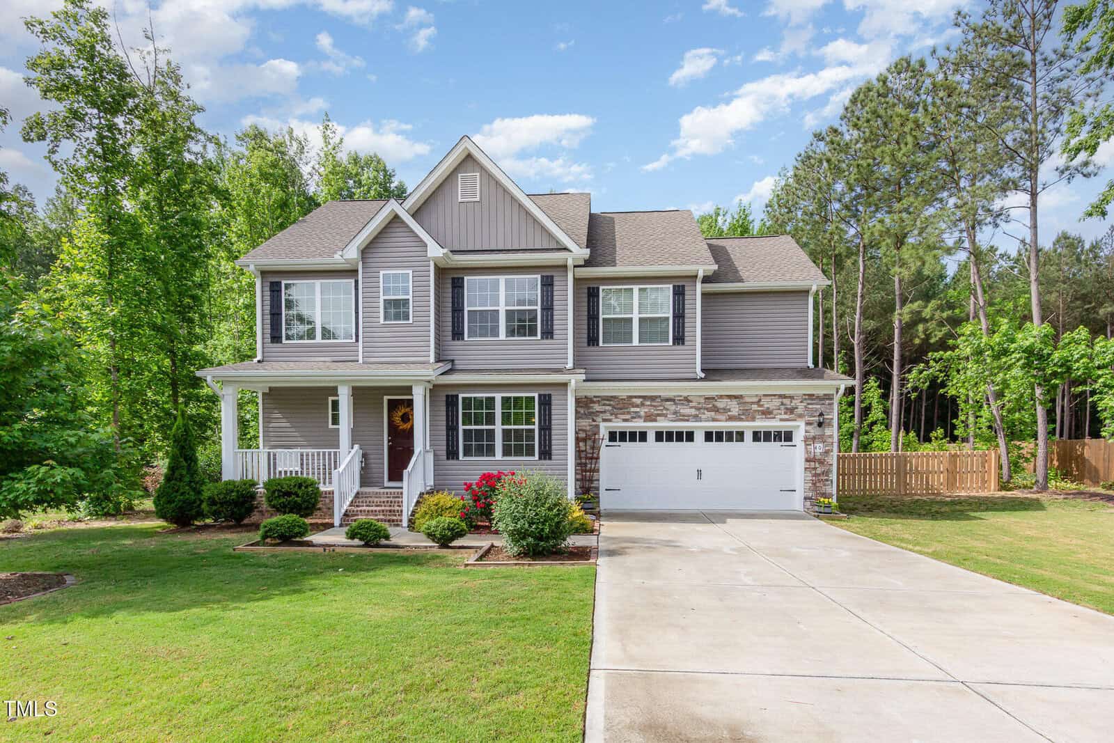Two-story suburban house with gray siding, stone accents, and a white two-car garage. The home has a front porch, green lawn, flower beds, and is surrounded by trees and a wooden fence.