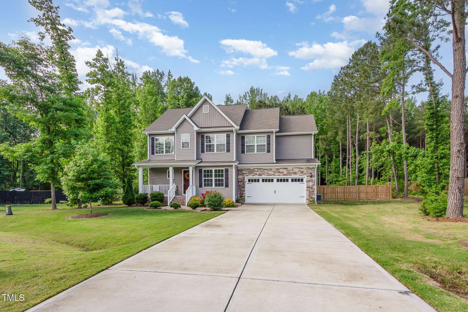 Two-story gray house with a white garage door and front porch, set back from a long concrete driveway. The home is surrounded by green grass, landscaping, and tall trees under a blue sky with clouds.