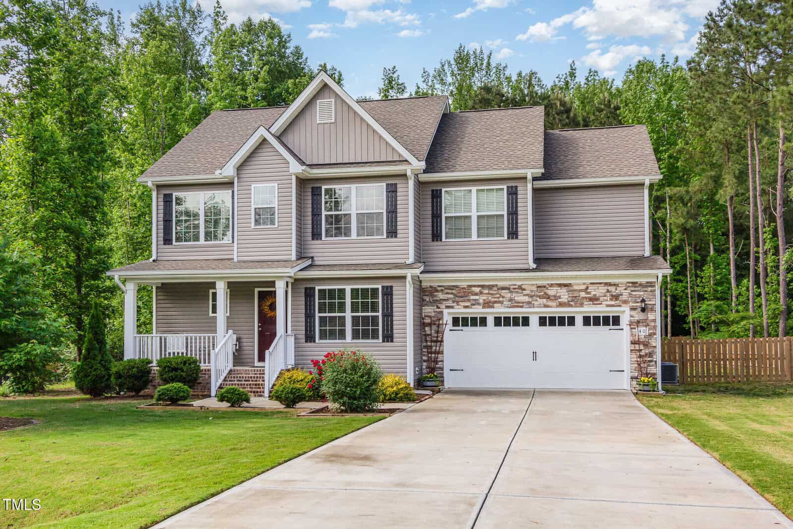 Two-story suburban house with beige siding, white trim, and stone accents. A covered front porch, attached two-car garage, and manicured lawn are visible, with trees and a wooden fence in the background.