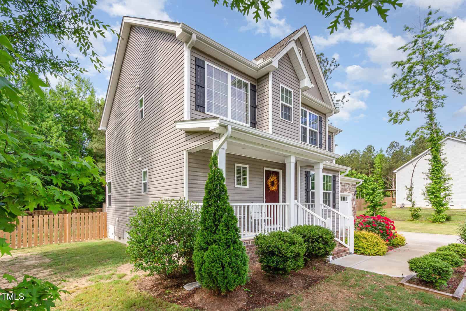 Two-story beige house with white trim, a small covered front porch, and a well-kept yard featuring shrubs, a tree, and blooming red flowers. A wooden fence and driveway are visible on the side.