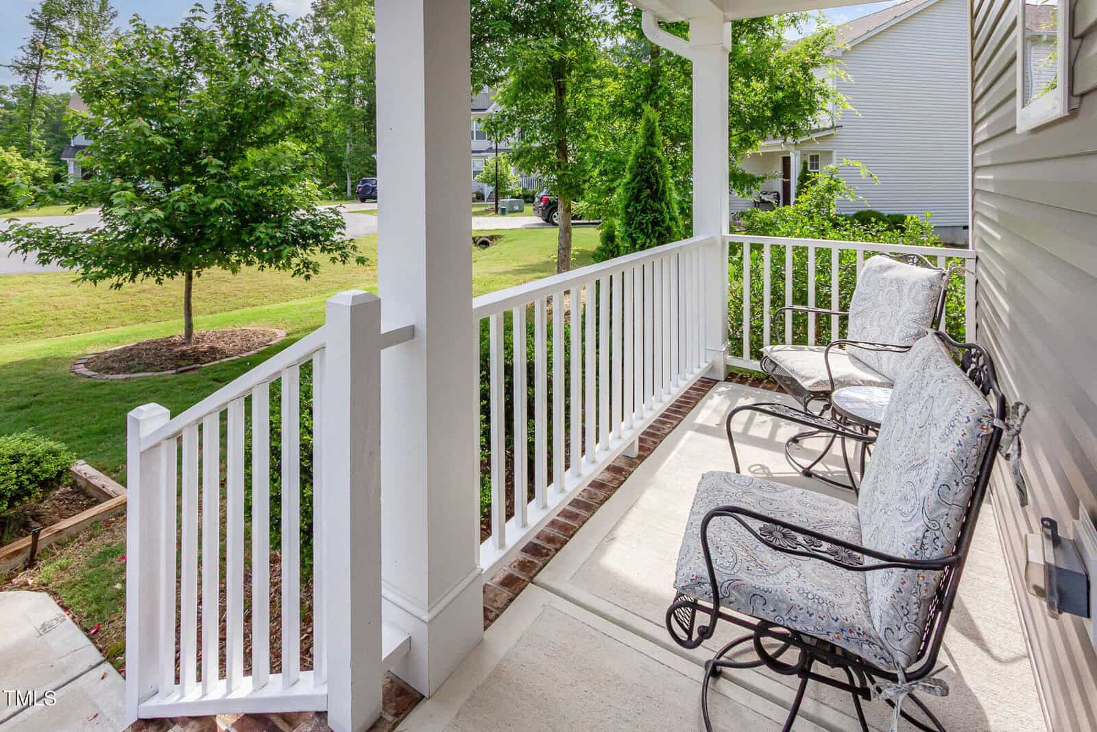 A covered front porch with white railings, two cushioned metal chairs, and a view of a green lawn and trees. The porch is attached to a light-colored house and overlooks a quiet residential street.