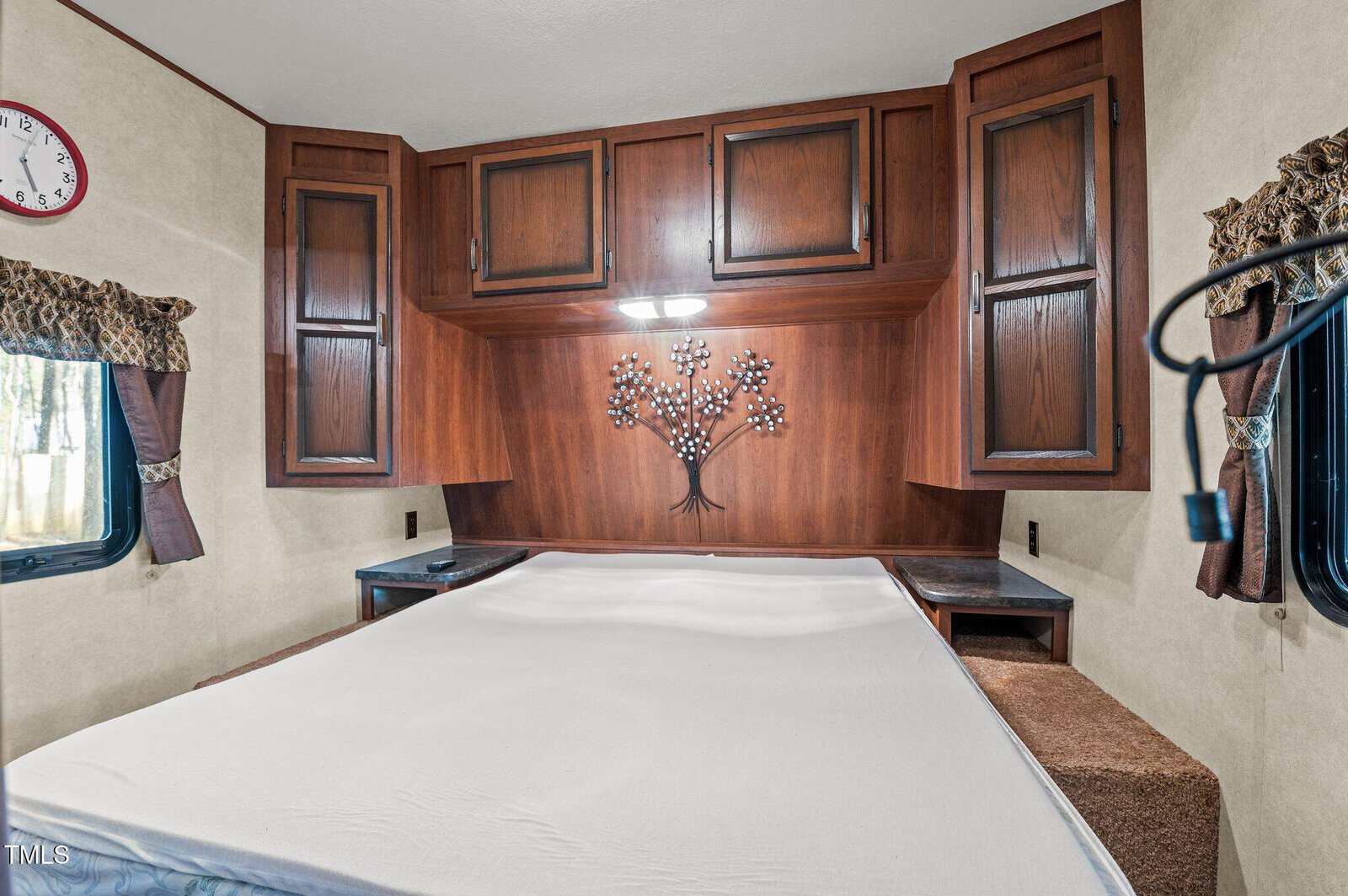 Bedroom inside a camper with a double bed, brown wooden cabinets above the bed, a metal tree wall decoration, two windows with curtains, a clock on the wall, and two small nightstands on either side of the bed.