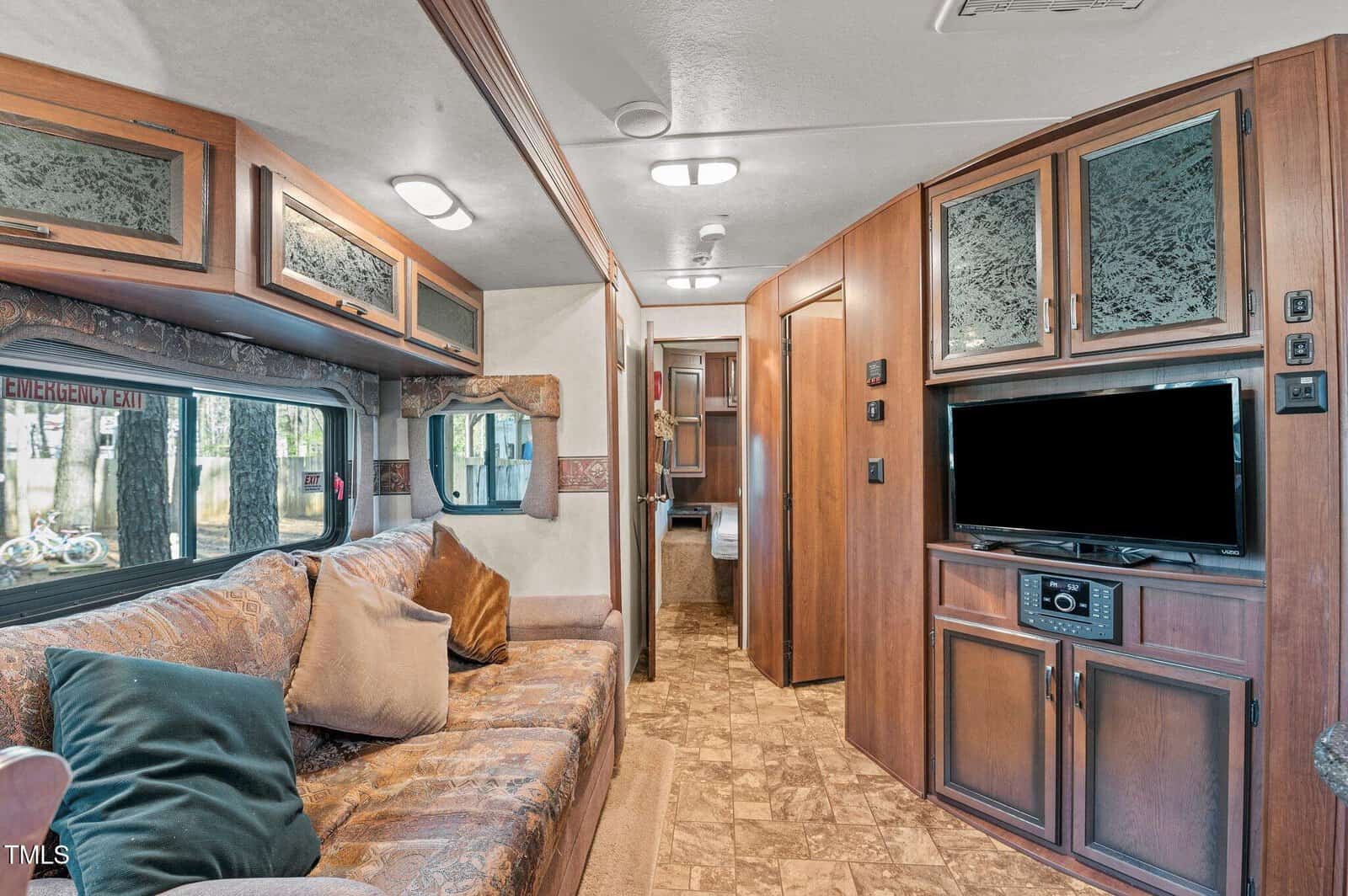 Interior of an RV with a patterned couch and multiple pillows on the left, wood cabinets and built-in TV on the right, and a narrow hallway leading to a bathroom in the background.
