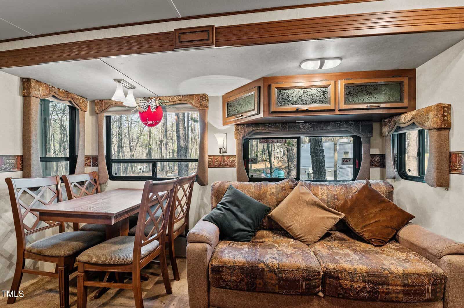 Dining area and living space inside an RV, featuring a wooden table with six chairs, a patterned sofa with three cushions, cabinetry above, and large windows showing a wooded outdoor view.