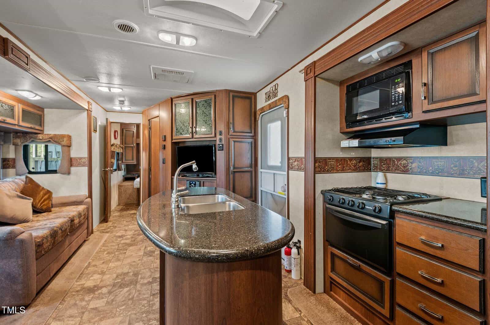 Interior of an RV kitchen and living area featuring a center island with a sink, stove, oven, microwave, wooden cabinets, small couch, wall-mounted TV, and neutral-toned flooring and walls.