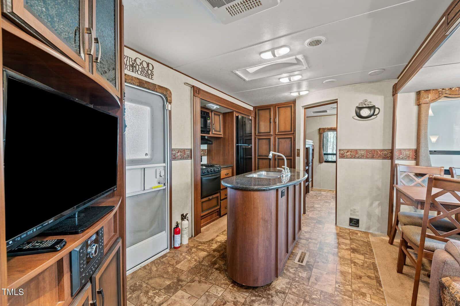 Interior of an RV kitchen with a central island, built-in appliances, wood cabinets, a dining area with chairs, wall-mounted TV, and a fire extinguisher near the door. The floor has a tile pattern.