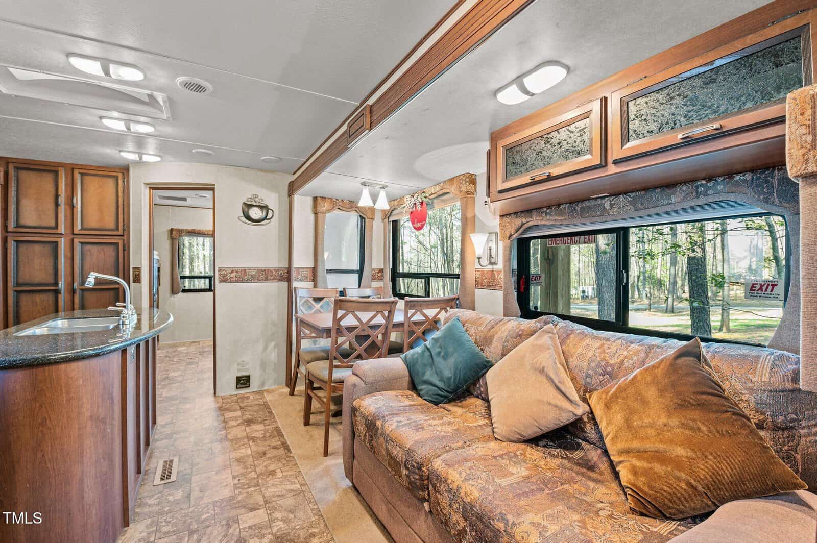 Interior of an RV featuring a brown patterned sofa with multiple pillows, a dining table with four chairs, wood cabinets, a curved kitchen counter with a sink, large windows, and view of trees outside.