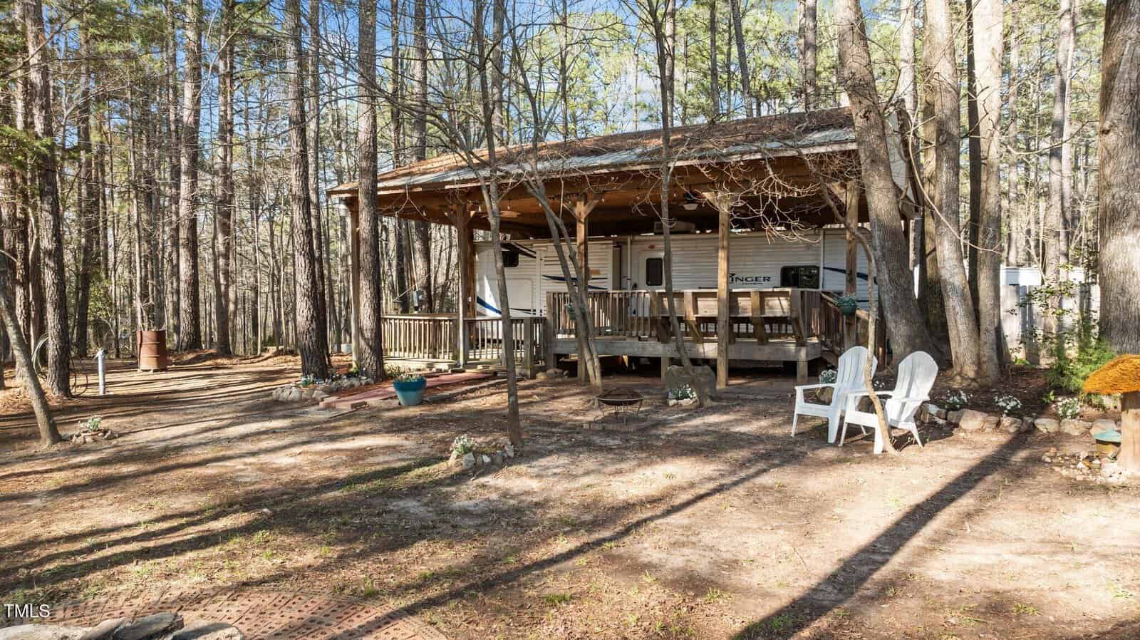 A covered wooden porch is attached to a camper trailer in a wooded area. Two white Adirondack chairs sit in the yard, and various potted plants and outdoor decor surround the porch. Tall trees fill the background.