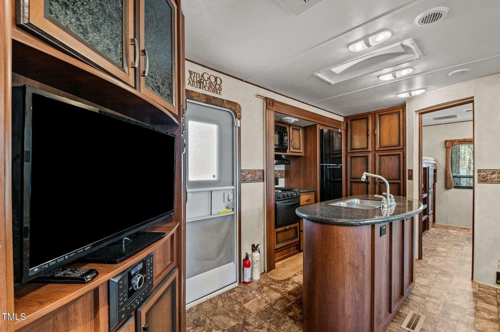 Interior of an RV kitchen showing wood cabinets, a black kitchen island with a sink, a wall-mounted TV, a stove, microwave, refrigerator, and a door. Fire extinguisher is near the door. Ceiling lights are visible.
